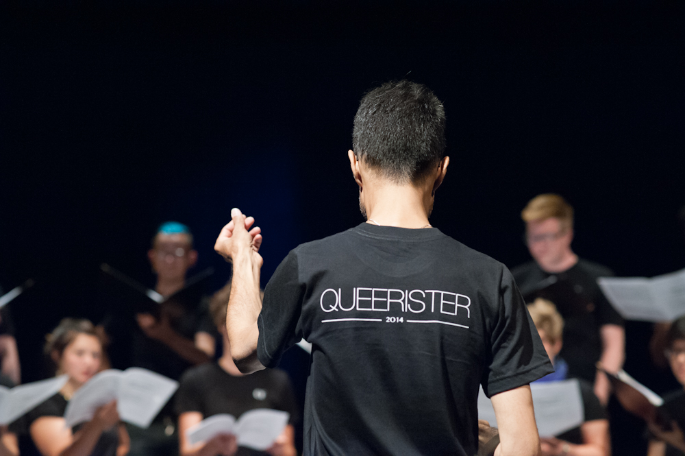 a person with brown skin and short black hair conducting a choir in a dark theatre