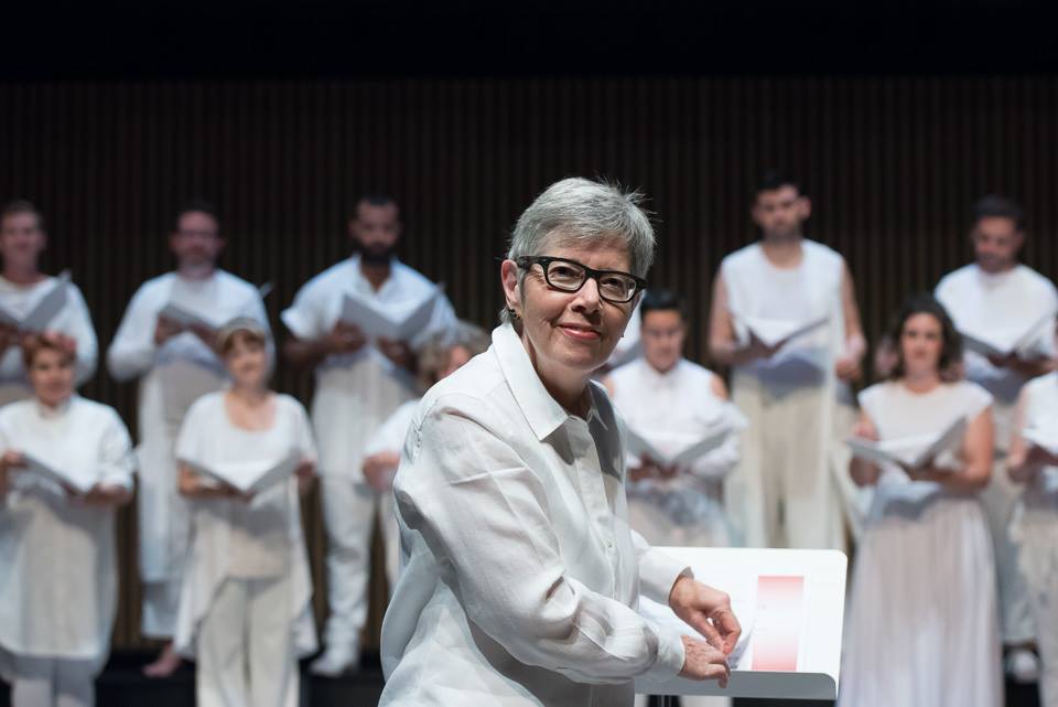 A person with short white hair turned and smiling, in front of a choir wearing all white