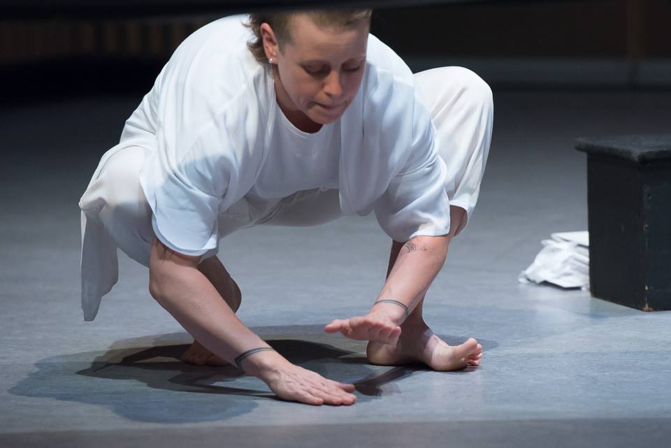 a barefooted singer wearing all white crouched onstage drumming on the floor